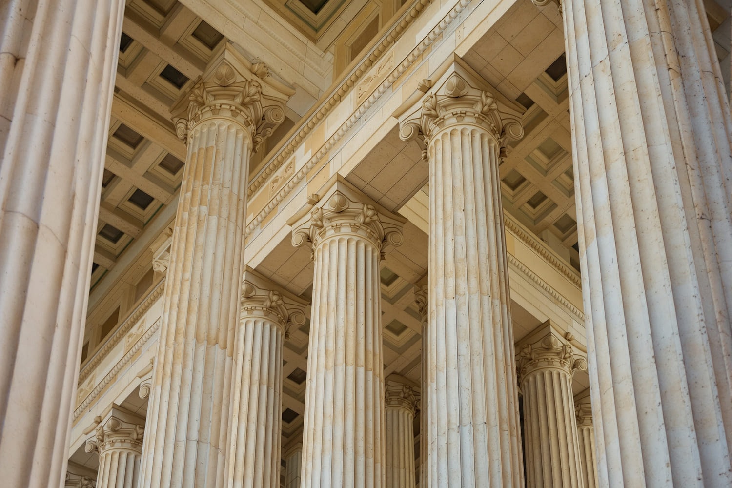 A row of stone columns beneath a coffered portico ceiling, photographed from below in morning light