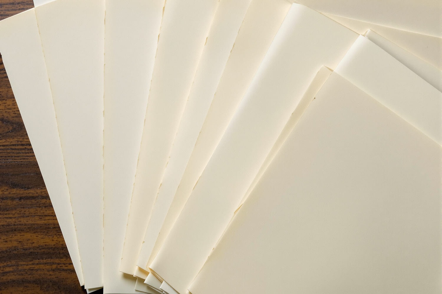 Ten blank sheets of heavy cream paper fanned out in an overlapping row on a dark wooden surface